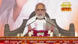 A man with a white beard and glasses speaks into two microphones, his hands gesturing slightly. The Aastha Bhajan logo is visible behind him.