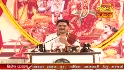 A man with a mustache, eyes closed, sits at a podium, holding a book. Behind him, a vibrant mural depicts Hindu deities.