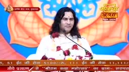 A man with long, dark hair speaks into a microphone, a garland of red flowers around his neck. He's on a stage with a large, stylized lotus flower behind him, likely for a broadcast on Aastha Bhajan.