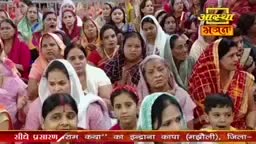 A crowd gathers, many women in colorful saris and headscarves, their faces turned towards something unseen. A banner at the bottom displays Hindi text, hinting at a religious broadcast.