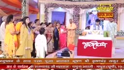 A man in a saffron robe stands near a red podium with Hindi script. A group of people in colorful Indian attire are gathered around, some looking towards the stage.