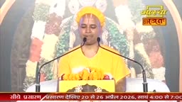 A man in a bright yellow robe and head covering speaks into two microphones. He stands before a backdrop of colorful garlands, likely for a devotional program.