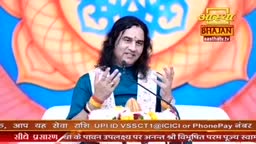A man with long dark hair speaks animatedly, gesturing with both hands. He's on a stage with a vibrant, lotus-like backdrop, likely for a broadcast on Aastha Bhajan.