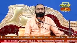 A man with a beard, wearing an orange kurta, speaks into a microphone. He sits on a decorative chair, a logo for Aastha Bhajan Live visible behind him.