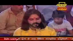 A man with long hair and a beard, wearing a yellow kurta, sits with his eyes closed. Beside him, a young boy in a white t-shirt looks down. The Aastha Bhajan logo is visible in the background.