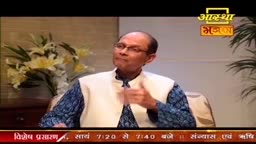 A man in a white vest over a blue patterned kurta speaks, gesturing with his right hand.  Behind him, a vase of white lilies sits on a dark wooden surface.