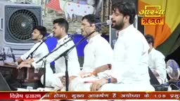 A group of men in white kurtas are seated, singing. One man is playing a harmonium while another holds a microphone.