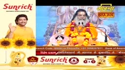 A man in a yellow shirt smiles and gestures with his hand. Behind him, a man with a long beard sits adorned with flower garlands.