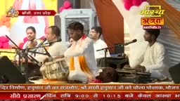 A musician plays the tabla, his hands a blur of motion over the drums. Another man, dressed in white and orange, sings into a microphone, his head bowed in concentration.