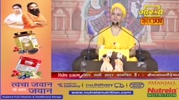 A speaker in a yellow robe addresses a crowd from behind a podium. To the left, advertisements display health products.