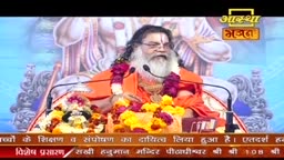A man with a long white beard, dressed in orange robes, sits on a decorated throne. He speaks into a microphone, surrounded by vibrant floral arrangements.