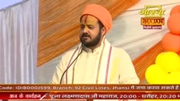 A man in an orange turban speaks into a microphone, his white tunic adorned with a striped sash. The Aastha Bhajan logo is visible behind him, along with clusters of orange and yellow balloons.