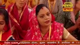 A woman in a bright pink sari speaks animatedly, her bindi prominent against her forehead. She sits among others, many also in vibrant Indian attire, with a yellow lanyard around her neck.