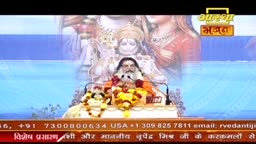 A man in orange robes sits before a backdrop of deities, speaking into a microphone. Garlands of yellow and white flowers adorn the podium in front of him.