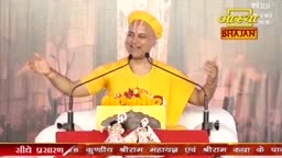 A man in a bright yellow robe and cap speaks with animated hands, his voice amplified by microphones. He stands before a podium adorned with flowers and small statues, likely during a spiritual discourse in India.