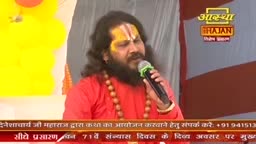 A man with a yellow and red mark on his forehead sings into a microphone. He wears a bright orange robe and a string of wooden beads.