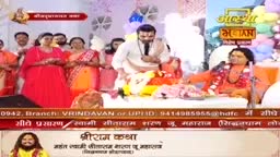 A man in a white kurta gestures with his hands while seated on a raised platform. To his left, women in vibrant saris stand, listening intently.