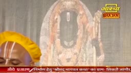 A man with a yellow cloth draped over his head and white markings on his forehead stands before a large, ornate depiction of a deity. The Aastha Bhajan logo is visible in the upper right corner.