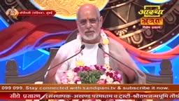 A man with a white beard and glasses speaks into two microphones, a garland of flowers around his neck. Behind him, a screen displays the Aastha Bhajan logo.
