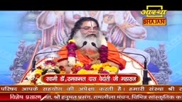 A man with a long white beard, wearing an orange robe, sits behind a podium adorned with flowers. Two microphones stand before him, and behind him, a large, stylized image of a deity is visible.