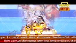 A man with a long beard, wearing a yellow robe and garlands, sits on a raised platform. Behind him, a large image of a deity is projected onto a screen.