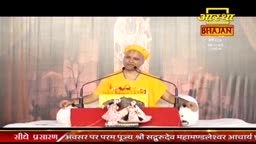 A man in a bright yellow kurta and turban speaks into a microphone. He sits behind a red podium adorned with small statues.