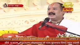 A man with a mustache and a red tilak on his forehead speaks into a microphone, his mouth open in mid-sentence. He wears a bright red robe over a white garment, and a garland of yellow flowers rests on the table before him.