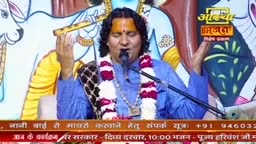 A man in a blue kurta sings into a microphone, his eyes closed. Garlands of white and maroon flowers drape around his neck.
