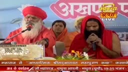 A man in a red turban and robes speaks into a microphone, his long white beard framing his face. Beside him, another man in a red hooded garment holds a microphone to his mouth.