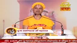 A man in a bright orange robe and headwrap speaks into two microphones. He's on a stage, a garland of orange flowers draped across the front of the podium before him. The Aastha Bhajan logo is visible in the upper right corner.