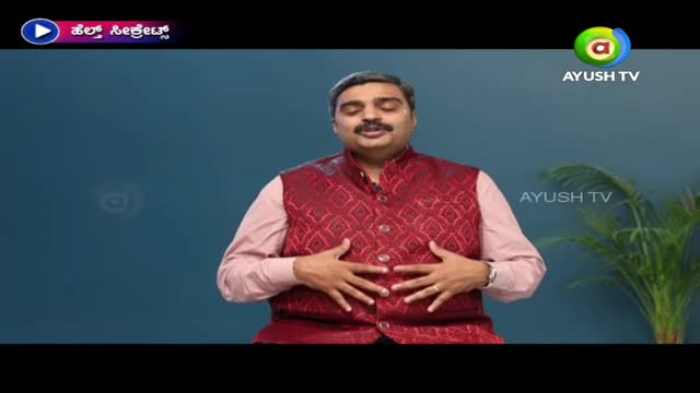 A man in a patterned red vest and pink shirt is on Ayush TV, gesturing with both hands over his stomach. He's speaking, and the studio set has a simple blue background with a small plant.
