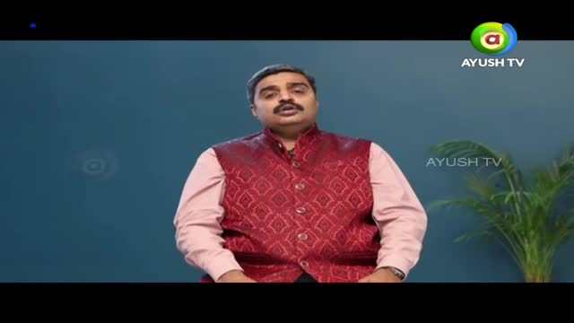 A man in a patterned red vest and pink shirt is speaking, likely on a program for Ayush TV in India. Behind him, a green plant sits to the right, and the Ayush TV logo appears in the upper right corner.
