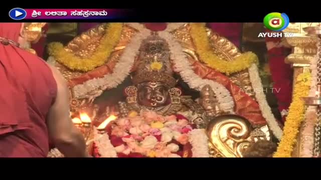 A priest in a red robe is performing a ritual in front of a decorated idol, with Ayush TV's logo visible in the corner. The scene is bathed in the warm glow of oil lamps and adorned with garlands.
