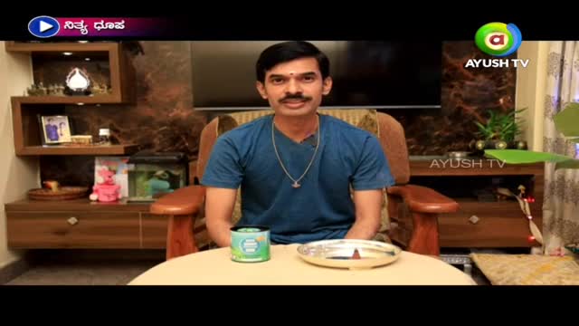A man in a blue shirt sits in a wooden chair, facing the camera. On the table in front of him are a silver plate and a small, green-labeled container, likely for a segment on Ayush TV in India.
