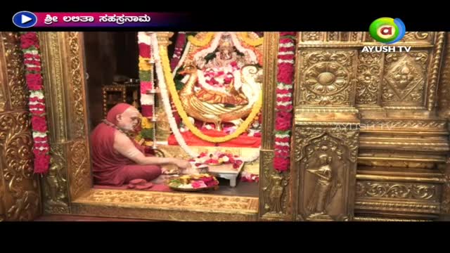 Inside a golden doorway, a robed man sits, arranging items on a small table. The scene is being broadcast on Ayush TV, with a decorated deity visible behind him.
