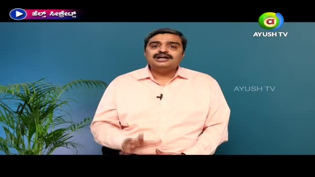 A man in a pink shirt is speaking, gesturing with his hands. The Ayush TV logo is visible, and a plant is in the corner.
