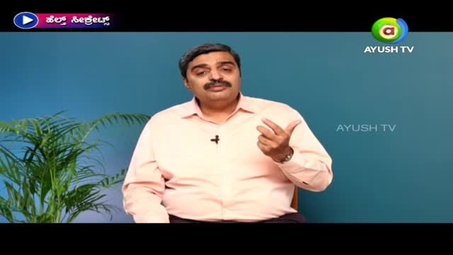 A man in a light pink shirt gestures with his right hand while speaking, framed by a blue background and a logo for Ayush TV. A small plant sits to his left, adding a touch of green to the scene.
