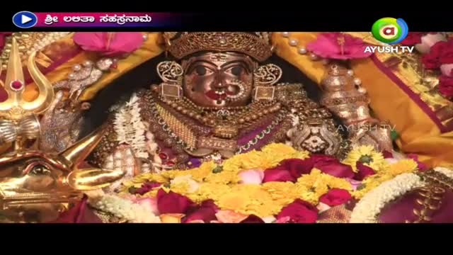 A jeweled deity, adorned with flowers, is the focus of a broadcast on Ayush TV from India. The camera is fixed on the elaborate face and ornate jewelry.
