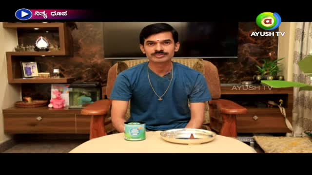 A man in a blue shirt sits in a wooden chair, facing the camera with a calm expression. A silver plate and a green tin sit on the table in front of him, while the Ayush TV logo is visible in the top right corner.
