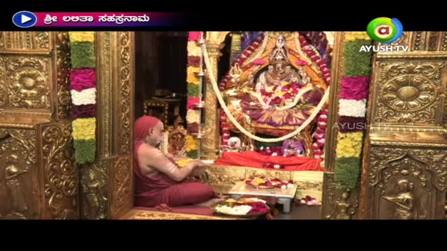 A robed man sits cross-legged, arranging flowers before a decorated deity. The scene is being broadcast on Ayush TV in India, framed by ornate, golden architecture.
