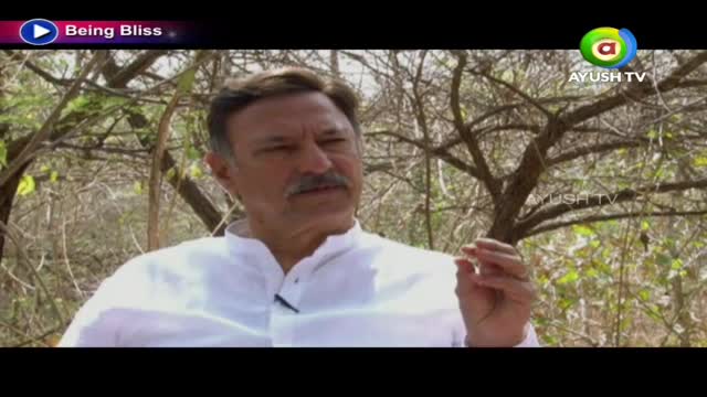 A man in a white shirt gestures with his right hand, speaking on Ayush TV. Behind him, bare tree branches create a natural backdrop for the interview.
