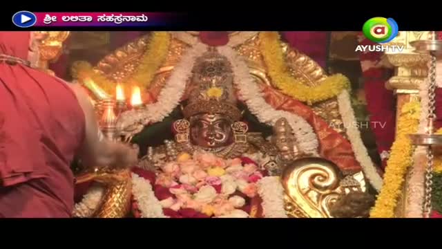 A priest in a red robe is holding a lit candle near a decorated idol, likely during a ritual. The scene is being broadcast on Ayush TV, with the title "Shree Lalitha Sahasranama" displayed in the corner.

