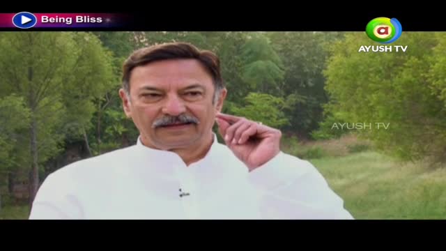 A man in a white shirt gestures near his ear, speaking on Ayush TV. Behind him, a blurred green landscape serves as the backdrop for the "Being Bliss" program.
