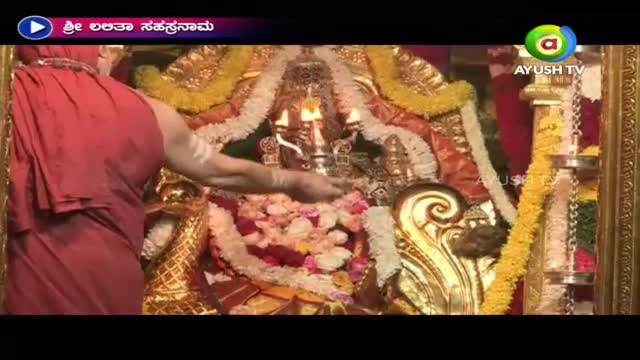 A robed figure, seen from the back, is reaching toward a shrine adorned with garlands and lit candles. It's a broadcast on Ayush TV, likely from India, capturing a religious ceremony.
