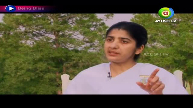 A woman in a white outfit gestures with her right hand while speaking, likely on Ayush TV. Behind her, a backdrop of green trees provides a natural setting for the interview.
