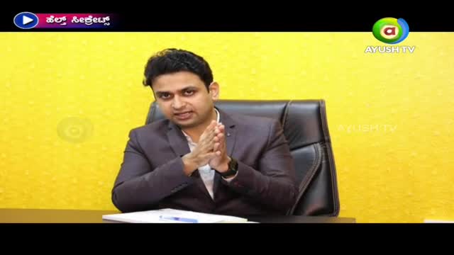 A man in a suit sits behind a desk, his hands clasped in front of him. The Ayush TV logo is visible in the corner, and a bright yellow backdrop fills the space behind him.
