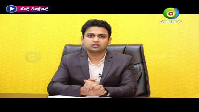 A man in a dark suit sits at a desk, speaking directly to the camera. The Ayush TV logo is visible in the corner, and the background is a bright yellow.
