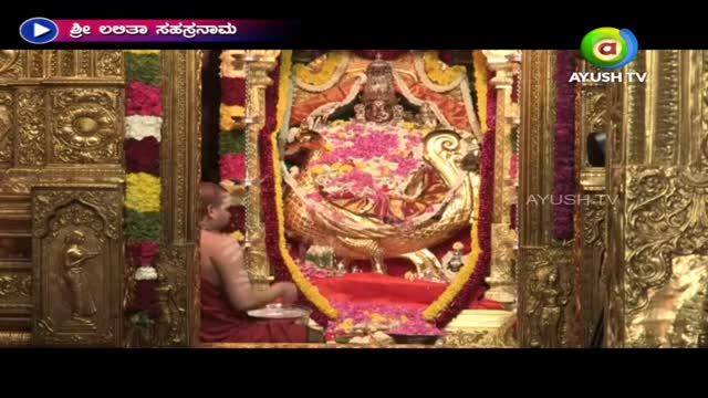 A robed figure is seated, holding a small plate, facing a golden shrine overflowing with flowers. The scene is broadcast on Ayush TV, with a title card in the top left corner.
