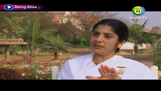 A woman in a white outfit gestures with her hands as she speaks, a logo for Ayush TV visible in the corner. Behind her, a garden with palm trees and blooming flowers fills the background.

