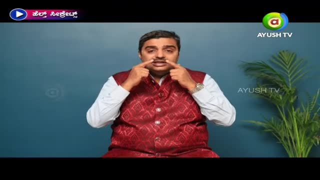 On Ayush TV, a man in a maroon vest is demonstrating something, his index fingers touching his nostrils. The background is a solid blue, and a plant sits off to the side.
